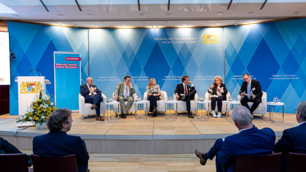 Sechs Personen diskutieren auf einem Podium miteinander. Im Hintergrund sind blaue Rauten sowie das Staatswappen zu erkennen.