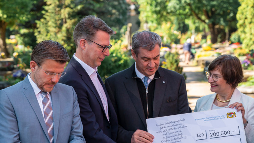 Nürnbergs Oberbürgermeister Marcus König, Wissenschaftsminister Markus Blume und Ministerpräsident Dr. Markus Söder (v.l.n.r.) übergeben einen Förderbescheid aus dem Entschädigungsfonds zur Unterstützung der Sanierung am St. Johannisfriedhof in Nürnberg.